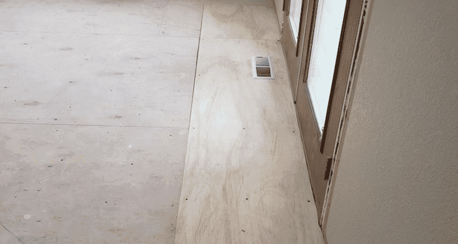 Interior view of unfinished floor with plywood and vent near large windows.