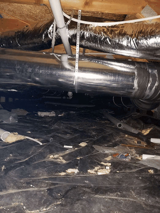 Ductwork and plumbing visible in a dark, cluttered crawl space under a house.