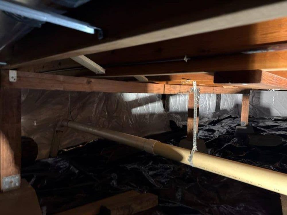 Underneath a house, wooden beams and plumbing visible on a dark plastic-covered floor.