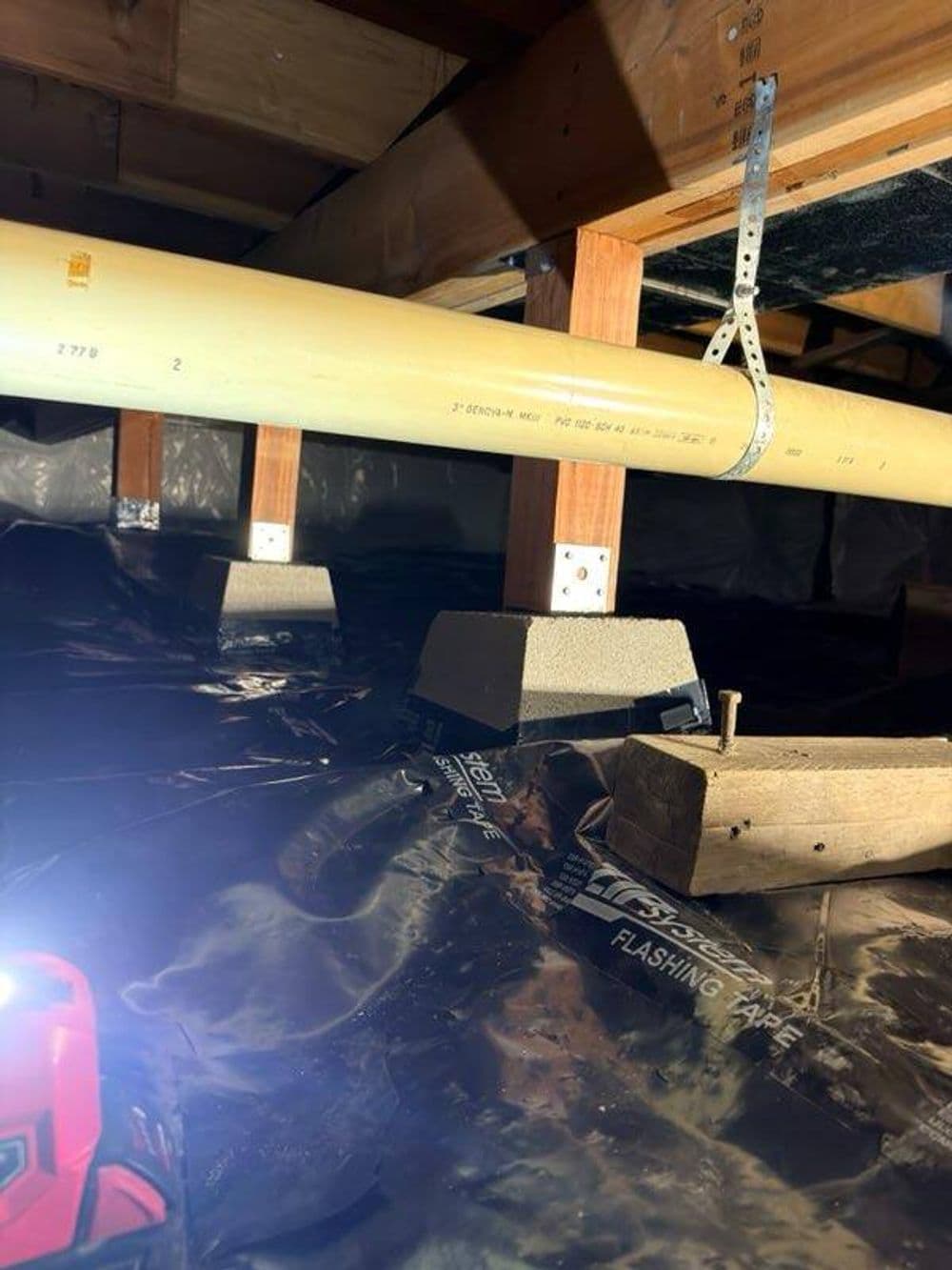 Pipes supported by mounts in a crawl space, illuminated by a flashlight.