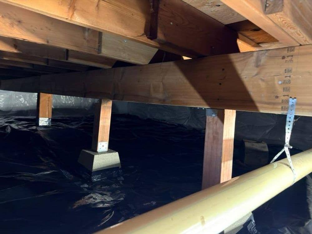 Wooden beams and supports in an encapsulated crawl space with black plastic lining.