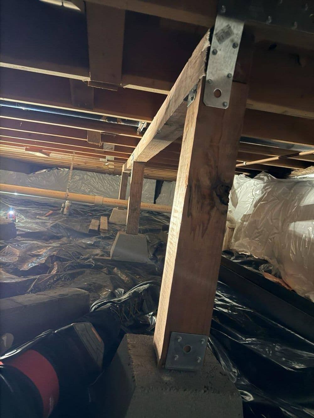 Wooden support beam in a crawl space, secured with metal brackets and resting on concrete blocks.