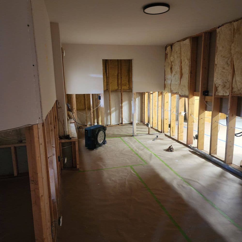 Renovation scene showing stripped interior walls, exposed framing, and plastic flooring.