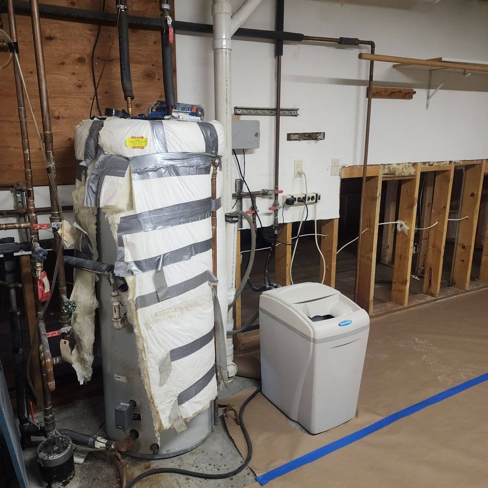 Water heater wrapped in insulation and tape, with a compact trash can nearby in a basement setting.
